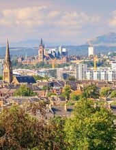 A viewpoint over the city of Glasgow from Queen's Park, taking in the greenery of trees with the city and Scottish landscape in the distance.